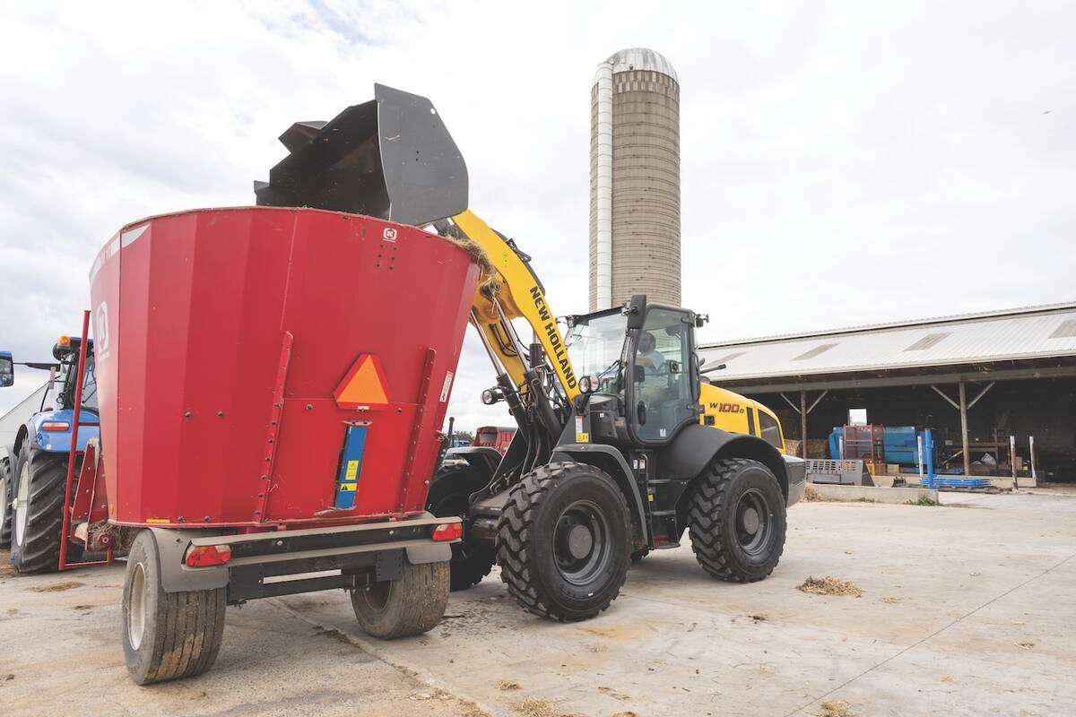 A New Holland W100D wheel loader dumps its bucket's contents over the lip of a large mixing tub very high off the ground.