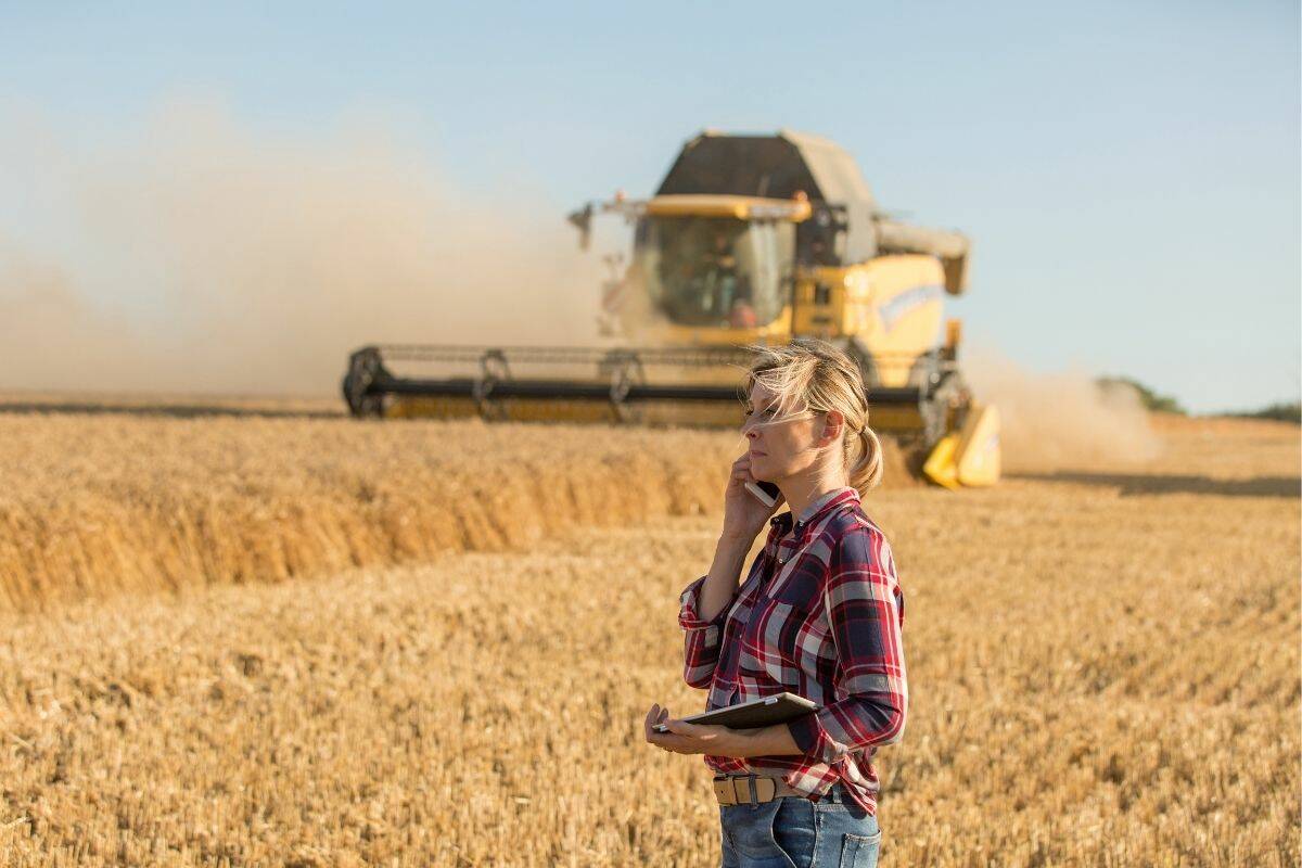 A woman talks on a smart phone while holding a tablet and standing in a field with a combine cutting a crop behind her.