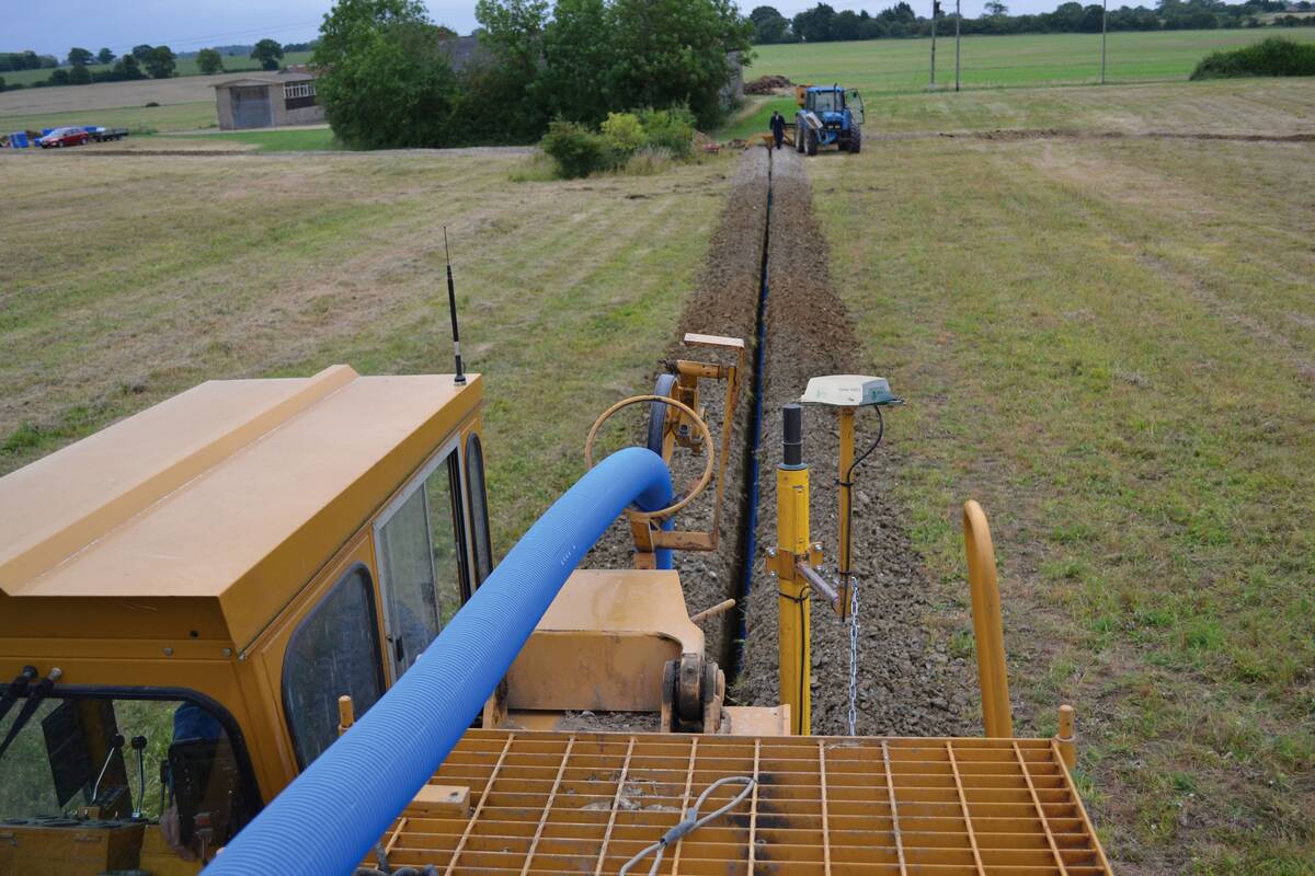 A trenching machine carves a trench into a field running blue plastic tile drainage piping into the trench simultaneously.