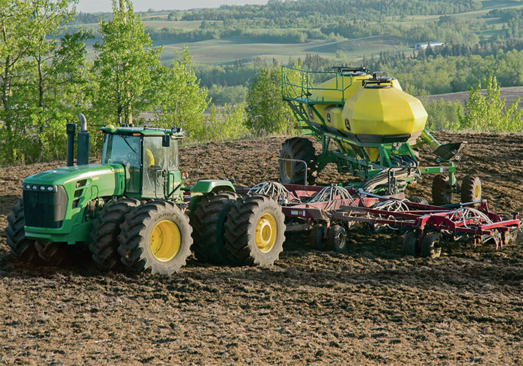 A large John Deere tractor with two big tires on each corner pulls a large air seeding rig in a field in a hilly area.