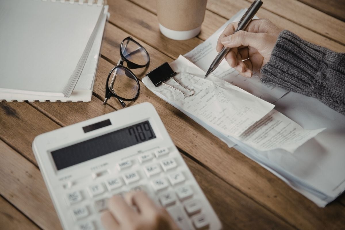 A woman's right hand holds a pen over some receipts on a table while her left hand enters numbers into a calculator.