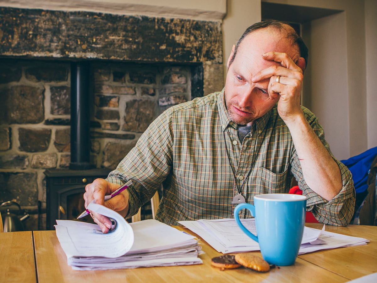 A man sits at a table with with one hand on his forehead and the other leafing through a pile of paperwork.