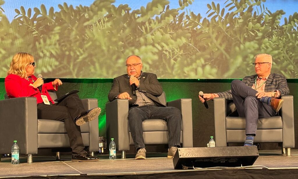 Three people, two men and a woman, sit in chairs on a stage at Canada