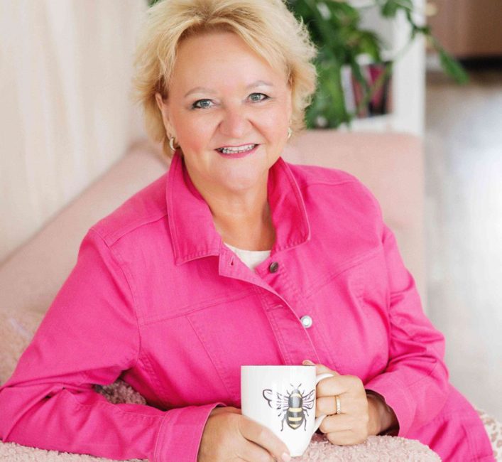 A woman in a brightpink top sits on a couch holding a coffee cup and smiling.