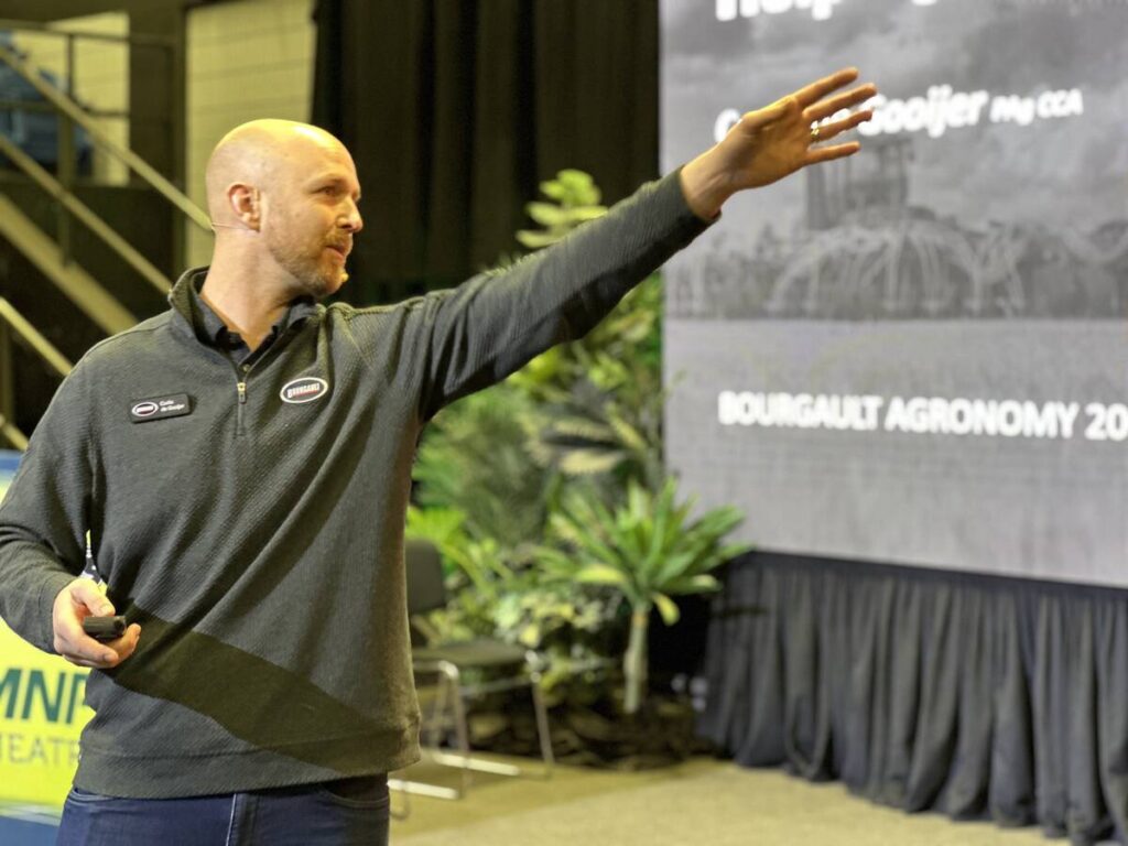 A bald man holding a "clicker" in his right hand gestures with his left toward an unseen screen he