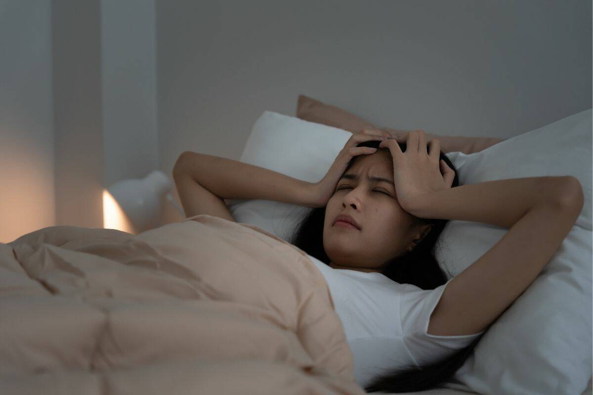 A woman laying in bed, holding her head with both hands, apparently having a bad night's sleep.