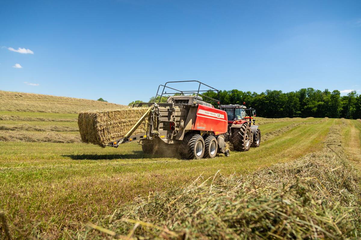 A tractor pulls a large square baler though a swathed field as it's expelling a bale.