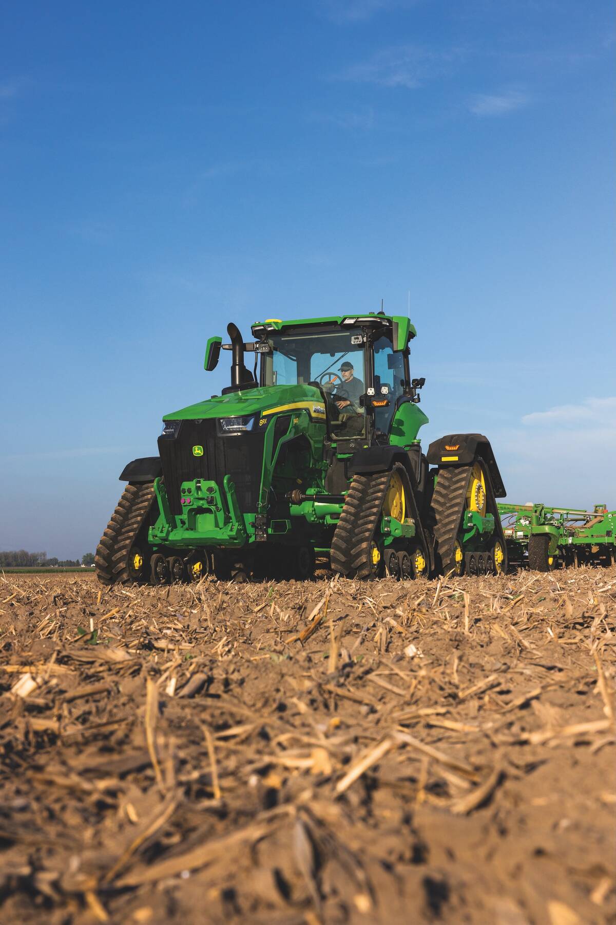 A large, tracked John Deere tractor pulls an implement through a field covered in litter.