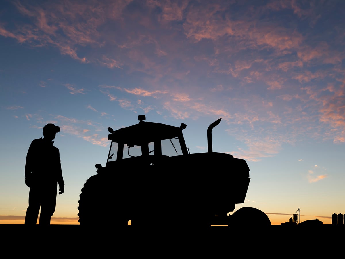 A farmer stands next to a tractor in a field, both perfectly silhouetted against the rising sun.