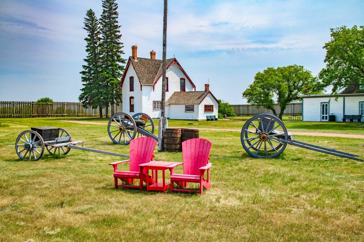 Three horse-drawn wagons and two red chairs sit on the lawn in front of two old houses at Fort Battleford historic site, Saskatchewan, Canada.