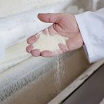The hand of a man wearing a white coat scoops some flour from a bin in a milling facility.