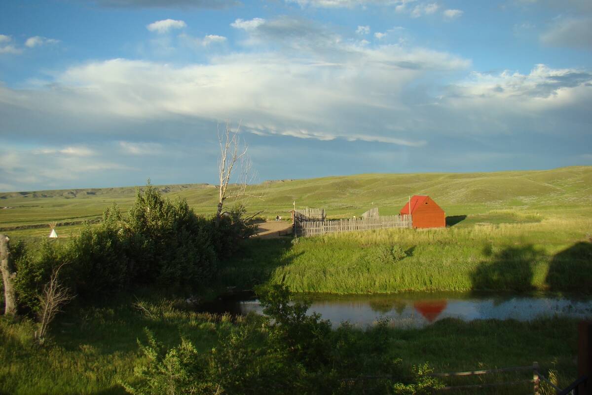 An old homestead freshly-painted red and some weathered gray fences contrast against a lush green landscape in summer as seen from a nearby hill with a small slough in between.
