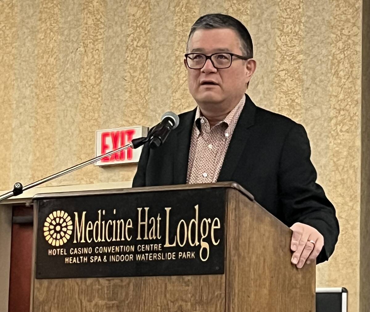 A man in glasses and a suit jacket speaks into a microphone at a lectern with "Medicine Hat Lodge" written on it.