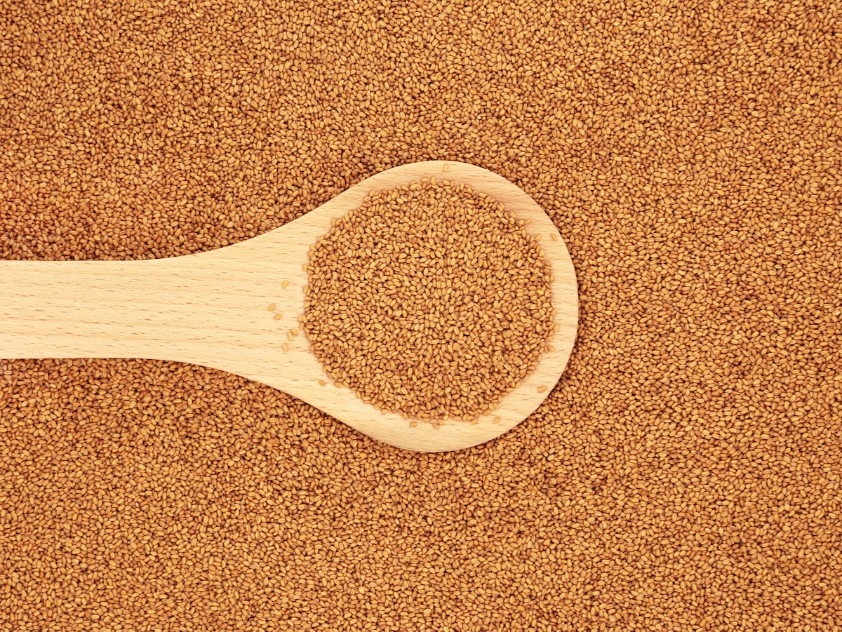 A wooden spoon full of camelina seed, set on a flat surface also covered in camelina seed.