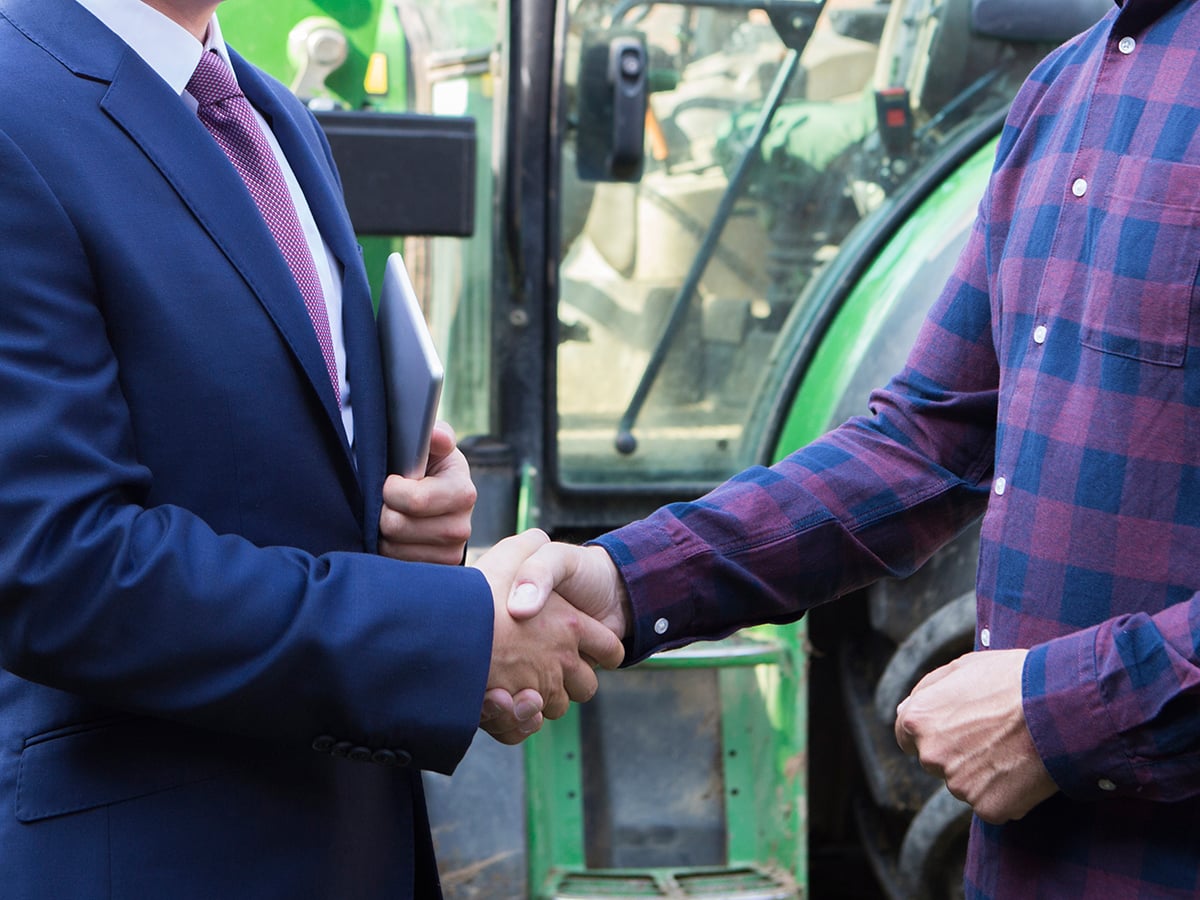 A farmer and a businessman in a suit shaking hands with a tractor in the b.ackground.