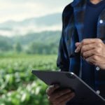 A farmer standing in a field with a tablet in hand and a barn blurred in the far background.