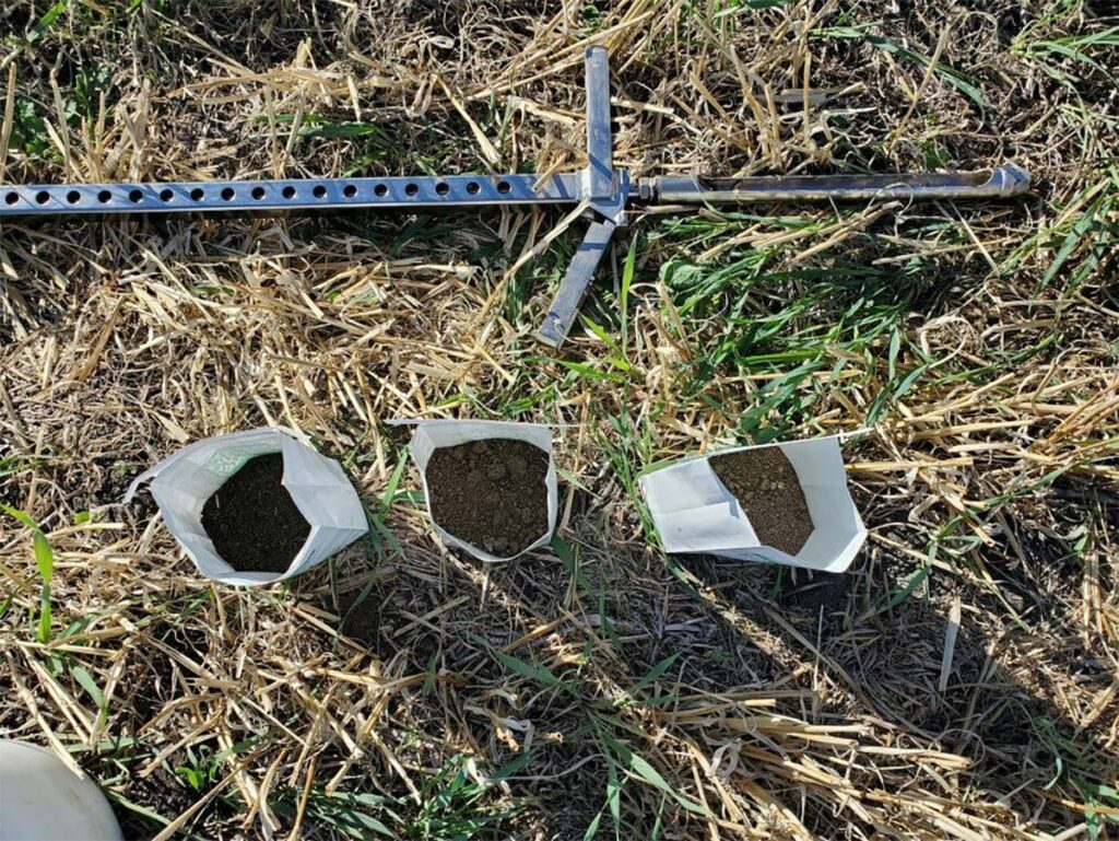 Three bags containing different-coloured soil samples sit on the ground next to a metal probe.