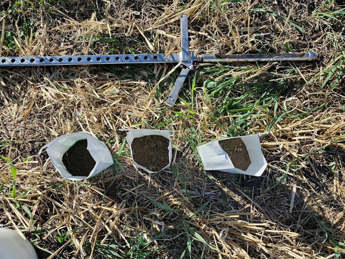 Three bags containing different-coloured soil samples sit on the ground next to a metal probe.