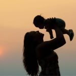 A mom holding up a baby silhouetted against a setting sun sky.