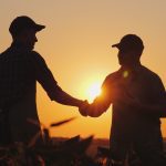 Two farmers wearing ball caps shaking hands in a field silhouetted against the setting sun.