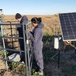 A woman and a man in coveralls work in a pasture next to a tripod with a box and what appears to be a rifle scope on top of it, surrounded by 3 corral panels with a large solar panel behind the woman.