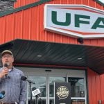 A man speaks into a microphone, and multiple media outlet microphones, outside a bright orange retail building during its grand opening. Sign overhead reads 'UFA'