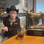 An elderly man in a black cowboy hat and vest leans on his cane while sitting in a bar with a drink in a glass and a large rodeo trophy featuring a Texas Longhorn bull on the table beside him.