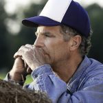 A stock photo of a farmer wearing a baseball cap, leaning on a large round bale, deep in thought.