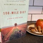 An apple and some figs are on a plate next to a book with the title, "The 100-Mile Diet" sitting on a counter in front of a white "subway tile" backsplash.
