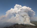 An aerial image of gray and white smoke rising high into an otherwise clear blue sky over a heavily-forested area below.