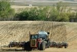 A tractor pulls an air seeder up a hill toward the camera. The furrows where the seed has just been placed as are some trees in the background.