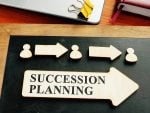 A wooden arrow with "Succession Planning" written on it sits on a desk next to a laptop and above it are two other, smaller arrows that point from the bust of one person to the bust of another and then to a third.
